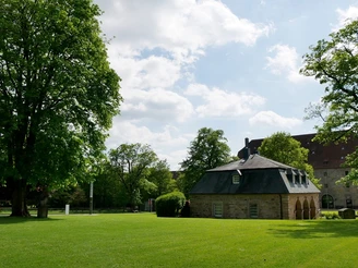 Ein kleines Steingebäude mit Schieferdach steht auf einer grünen Wiese, umgeben von Bäumen.