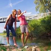 Nethemündung Zwei Frauen stehen barfuß in einem flachen Fluss und schauen lächelnd in die Ferne, im Hintergrund eine Brücke.