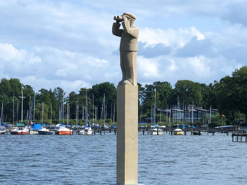 "Der Hafenmeister" Skulpturenpromenade Die Skulptur „Der Hafenmeister“ steht auf einem hohen Sockel am Wasser, umgeben von Segelbooten.