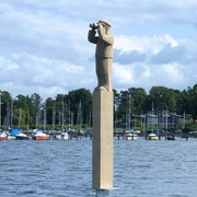 "Der Hafenmeister" Skulpturenpromenade Die Skulptur „Der Hafenmeister“ steht auf einem hohen Sockel am Wasser, umgeben von Segelbooten.