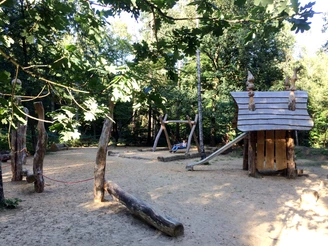 Kinderspielplatz am Waldlehrpfad Vosspäddken im Osnabrücker Land Spielplatz mit Holzhütte, Klettergerüst und Schaukel unter Bäumen im Sonnenschein.