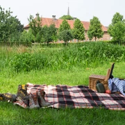 Das Kloster Clarholz beim Picknick im Blick Picknickdecke mit Korb und Schuhen auf ländlicher Wiese, im Hintergrund Bäume und ein Gebäude.