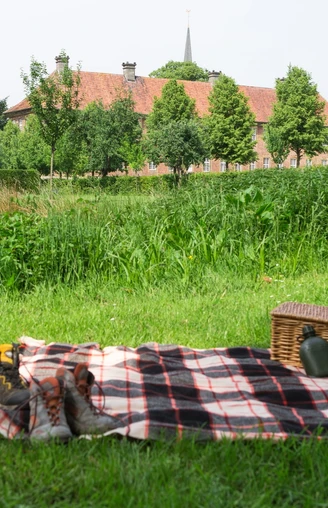 Das Kloster Clarholz beim Picknick im Blick Picknickdecke mit Korb und Schuhen auf ländlicher Wiese, im Hintergrund Bäume und ein Gebäude.