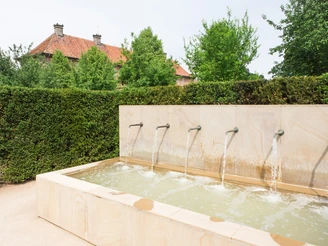 Brunnen im "Labyrinthgarten No.10" am Kloster Clarholz Ein moderner Brunnen mit sechs Wasserausläufen vor einer Hecke und einem roten Ziegelgebäude.