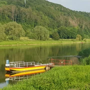 7_hx_fhu_faehrewehrden_stephanberg Eine gelbe Fähre mit rotem Geländer liegt ruhig an einem grünen Flussufer, umgeben von Wäldern.