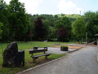 Kurpark mit Spielplatz <p>Ein grüner Park mit Bänken, Spielplatz, Bäumen und einem Kiesweg in einer ländlichen Umgebung.</p>