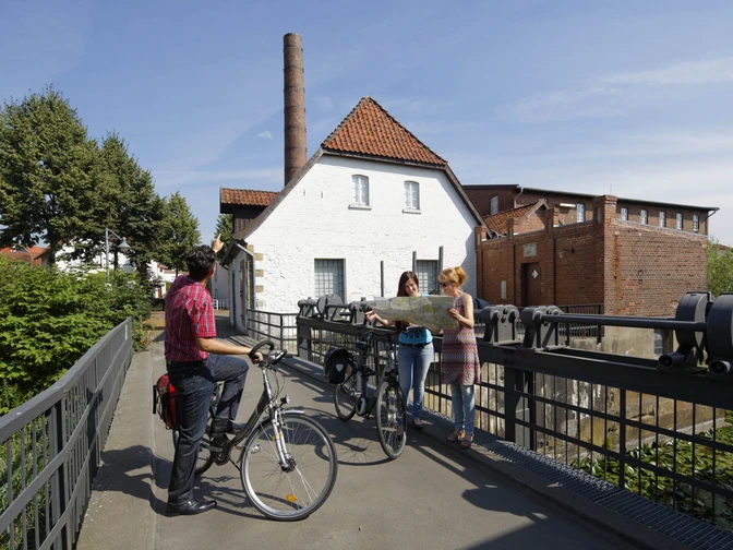 Radwanderer vor dem Tuchmacher Museum in Bramsche  Drei Radfahrer studieren eine Karte vor einer historischen Wassermühle an einem sonnigen Tag.