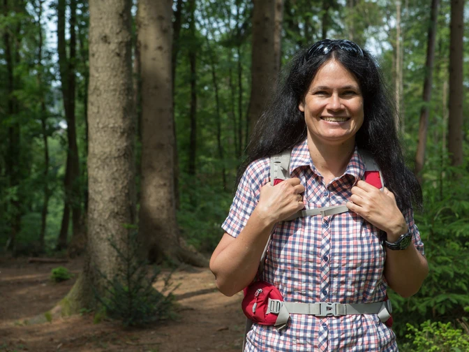Unterwegs im Wald Frau mit Rucksack lächelt im sommerlichen Wald vor hohen Bäumen und grünem Laub.