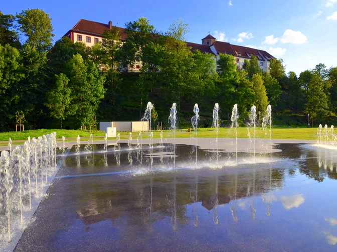 Springbrunnen vor einem weitläufigen, historischen Gebäude mit rotem Dach und grüner Umgebung.