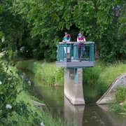 Bifurkation  Zwei Wanderer stehen auf einer Aussichtsplattform über einem kleinen Fluss im Grünen.