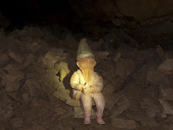Ein Hüggelzwerg Ein Zwerg mit Spitzhut sitzt auf Steinbrocken in einer dunklen Höhle.