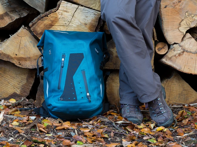 Wanderrast Ein blauer Wanderrucksack und Beine einer Person stehen auf buntem Herbstlaub vor Holzstapeln.