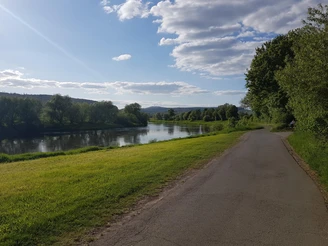 1_weseraue_abzweigholzminden_hx2_stephanberg Ein ruhiger Fluss schlängelt sich durch eine grüne Landschaft, flankiert von Bäumen und einem blauen Himmel.
