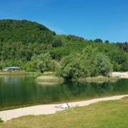 Grüne Hügel und ein klarer See mit Sandstrand, umgeben von Bäumen unter blauem Himmel.