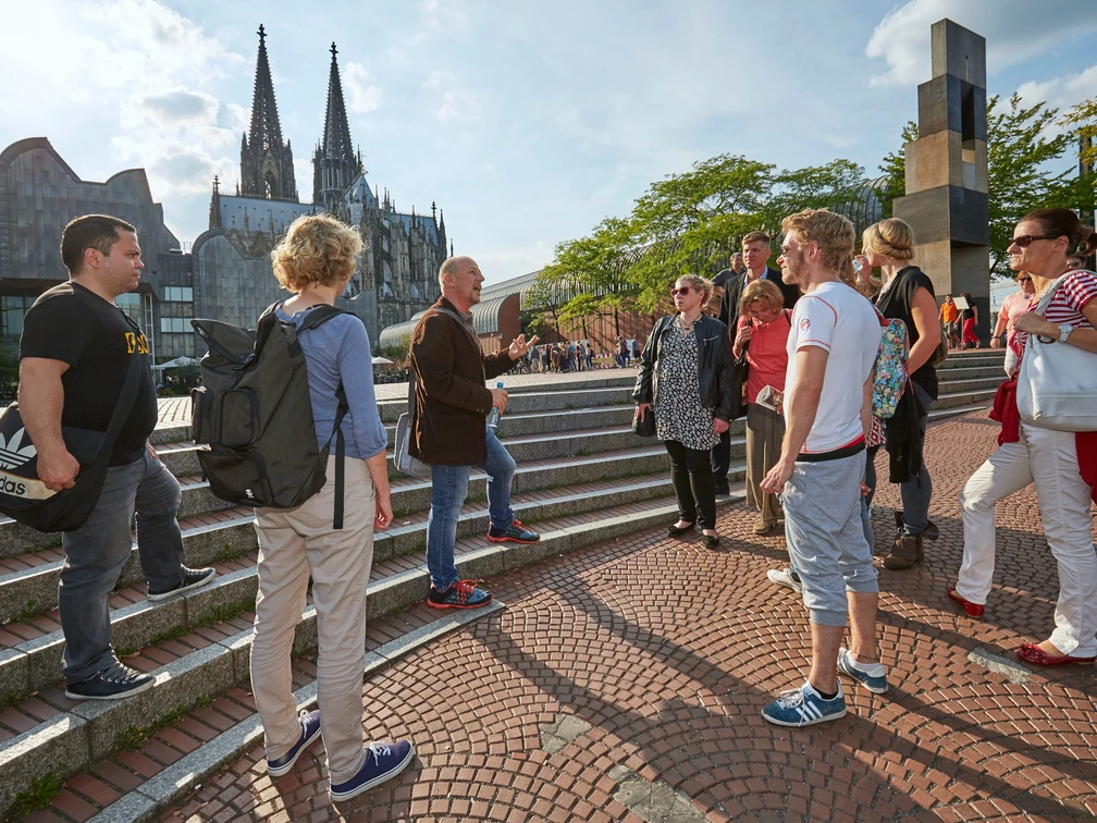 Eine Gruppe von Menschen steht auf einer Treppe in Köln. Im Hintergrund sind der markante Kölner Dom und moderne Architektur zu sehen. Ein Reiseleiter gestikuliert während einer Stadtführung im Sonnenschein, umgeben von interessierten Tourist*innen, die die Geschichte und Kultur Kölns erkunden.A group of people stand on a staircase in Cologne. The striking Cologne Cathedral and modern architecture can be seen in the background. A tour guide gestures during a city tour in the sunshine, surrounded by interested tourists exploring the history and culture of Cologne.