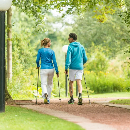 In Bewegung bleiben mit Nordic Walking  Zwei Personen in sportlicher Kleidung walken mit Stöcken auf einem Parkweg, umgeben von Bäumen.