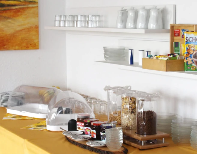 pension-zurweser-fruehstueck Frühstücksbuffet mit Cerealien, Marmelade und Saft, vor warmen Erdfarben und schlichtem Geschirr.