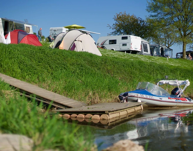 wesercamping-bootsanleger Campingplatz mit Zelten und Wohnwagen am Fluss. Ein Boot ist an einem Steg im Wasser vertäut.