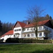 Naturfreundehaus Teutoburg Großes, modernes Gebäude mit rotem Dach, umgeben von Wald und Wiese an einem sonnigen Tag.