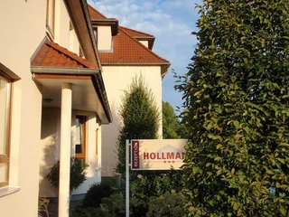 Seitenansicht Hotel Hollmann Hotel Hollmann mit rotem Ziegeldach und Empfangsschild, umgeben von grünen Büschen und Bäumen.
