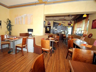 Cafe/Bäckerei Pension Mertens Innenansicht einer hellen Bäckerei mit Holzmöbeln, runden Tischen und großen Fenstern im Hintergrund.