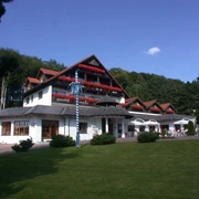 Aussenansicht Hotel Mügge am Iberg***, Oerlinghausen Dreigeschossiges Hotel mit roten Balkongeranien vor bewaldetem Hügel, blauer Himmel im Hintergrund.