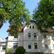 haus-kehne_07-05-2013 Das zweistöckige Haus mit mansardartigem Dach liegt zwischen grünen Bäumen, umgeben von blauen Himmel.