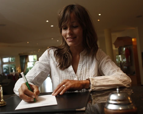 platzhalterbild_uebernachtung_twt_t-conrad Eine Frau schreibt in einer Hotelrezeption lächelnd auf Papier, umgeben von eleganter Einrichtung.