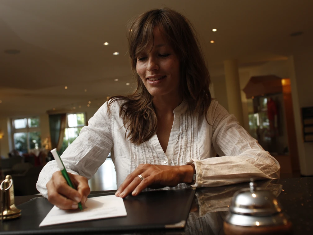 platzhalterbild_uebernachtung_twt_t-conrad Eine Frau füllt ein Formular an einem Hotelrezeptionstresen aus; daneben eine Glocke.