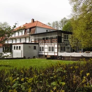 aussenansicht_haus-varlemann_warburg Fachwerkhaus mit rotem Ziegeldach, umgeben von Bäumen und Hecken, mit gepflegtem Rasen im Vordergrund.