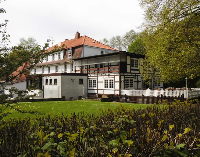 aussenansicht_haus-varlemann_warburg Fachwerkhaus mit rotem Ziegeldach, umgeben von Bäumen und Hecken, mit gepflegtem Rasen im Vordergrund.
