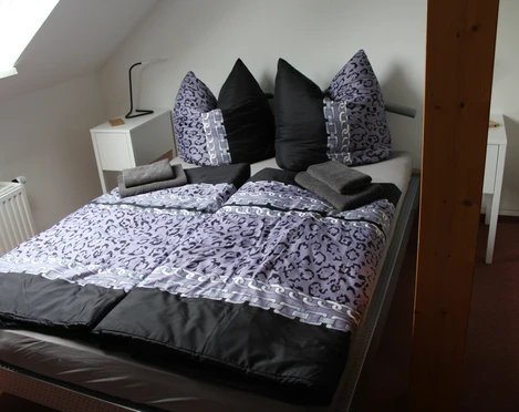 Ein einladendes Schlafzimmer mit einem großen Bett in dunklen und hellen Lilatönen, verziert mit flauschigen Kissen, angrenzend an eine obere Etage mit Dachschräge.