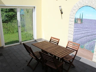 Eine Terrasse mit Holzmobiliar, einer großen Fensterfront und einer Wandmalerei im mediterranen Stil.