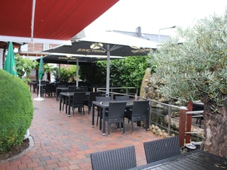 Geräumige Terrasse mit schwarzen Stühlen und Tischen unter weißen Sonnenschirmen und grüner Kulisse.