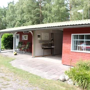 Ein rotes Holzgebäude in einer bewaldeten Umgebung mit überdachtem Sitzbereich und Grünflächen.