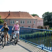 Zwei Frauen auf Fahrrädern überqueren eine Brücke vor dem historischen Grafenschloss in Hoya.