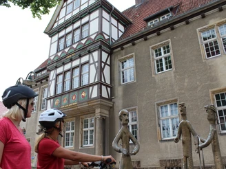 Altes Amtsgericht Zwei Radfahrerinnen mit Helmen betrachten moderne Bronzefiguren vor einem Fachwerkgebäude.