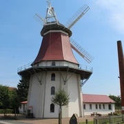 Gallerieholländerwindmühle Emtinghausen Historische Windmühle in Emtinghausen, mit rotem Dach und Flügeln, umgeben von Bäumen und Gebäuden.