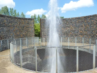 Wasserfontäne bricht aus rundem, metallumzäuntem Krater hervor, von hohen Steinwänden umgeben.