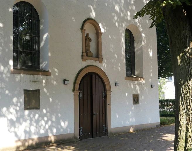 Eingang der weißen Pfarrkirche St. Joseph in Paderborn-Marienloh mit brauner Holztür unter Bäumen.