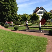 Menschen trainieren an Fitnessgeräten im Freien, umgeben von grüner Wiese und Bäumen, vor einem Haus.