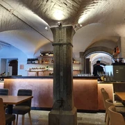 Beleuchtetes Kellergewölbe, eine Säule in der Raummitte und mehrere Tische mit Stühlen. An der hinteren Wand ist eine beleuchtete Bar mit verschiedenen Flaschen.