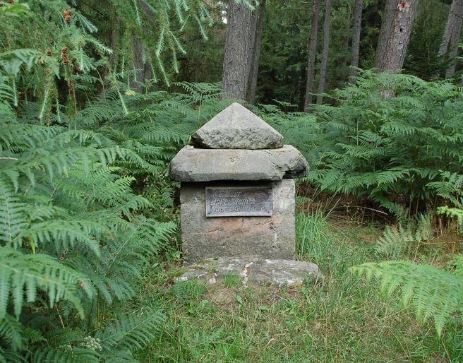 Kleiner Herrgott Steinernes Marterl mit pyramidenförmigem Dach, umgeben von Farn und Nadelbäumen im Wald.