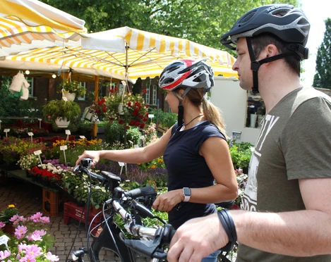 Achimer Wochenmarkt Zwei Radfahrer mit Helmen betrachten einen Marktstand voller bunter Blumen auf einem Wochenmarkt.