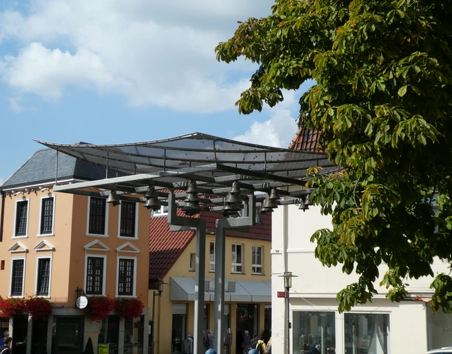 Achimer Glockenspiel Eine moderne Glockenspielinstallation vor historischen Gebäuden mit Blätterdach im Vordergrund.