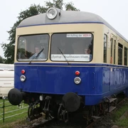 Kaffkieker Ein blauer nostalgischer Zug auf Gleisen, umgeben von grüner Landschaft und einem Wohnwagen.