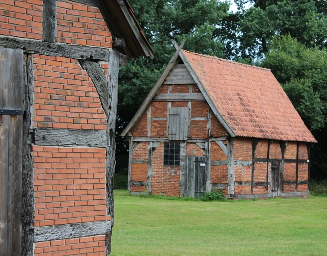 Schafstallviertel Hülsen Zwei historische Fachwerkgebäude mit Backsteinwänden stehen auf einer grünen Wiese vor Bäumen.