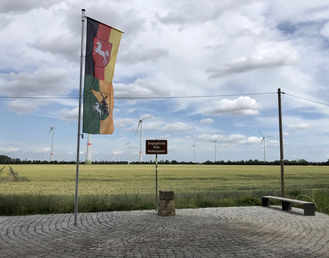 Geografischer Mittelpunkt Niedersachsens (16).jpg Vor einem windkraftbestückten Getreidefeld weht die Flagge Niedersachsens neben einem Wegweiser.