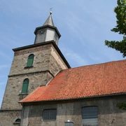 Martinskirche Hoya Alte Kirche mit rotem Ziegeldach und Turm mit Uhr unter einem blauen Himmel, umgeben von Bäumen.