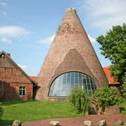 Großes, rundes Backsteingebäude der historischen Glashütte Gernheim mit Holzschindeldach und Fensterfront.